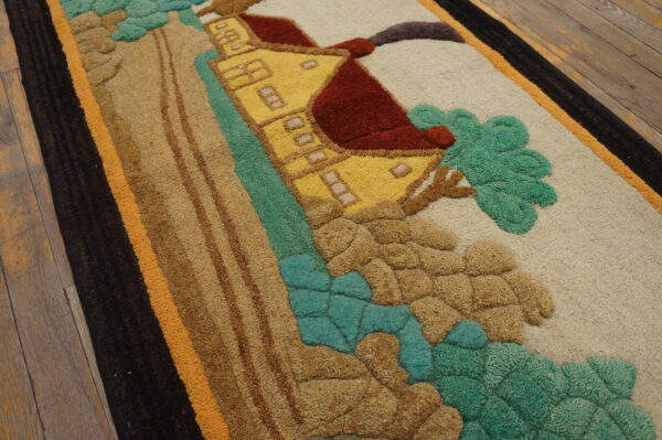 Close-up of a sculpted rug depicting a yellow house and green landscape, bordered in black and orange on wood flooring.