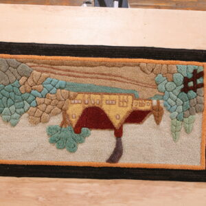 Textured rug showing a stylized house and teal trees against a brown landscape, framed by black and orange borders.