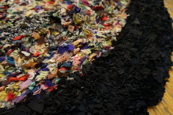 Highly textured rug with multicolored fabric strips adjacent to solid black strips on a wooden floor.