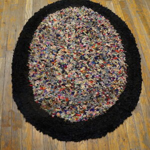 A high-pile oval rug with a multicolored rag center and thick black border on plank wood flooring.