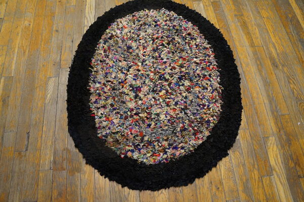 A high-pile oval rug with a multicolored rag center and thick black border on plank wood flooring.