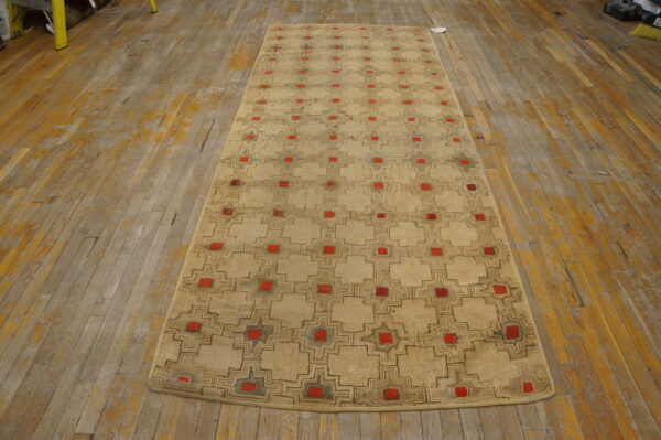 Beige runner rug with brown outline geometric pattern and bright red squares on distressed wood floor.