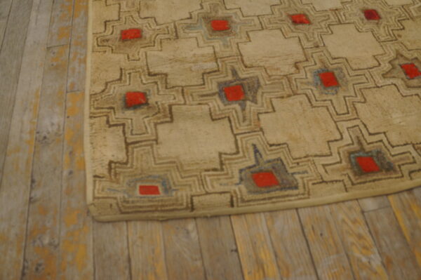 Beige rug with a repeating brown, blue, and red geometric motif resting on worn wood plank flooring.