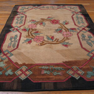 Tan rug with pink floral wreath, dark brown frame, and red-teal lattice border on hardwood flooring.