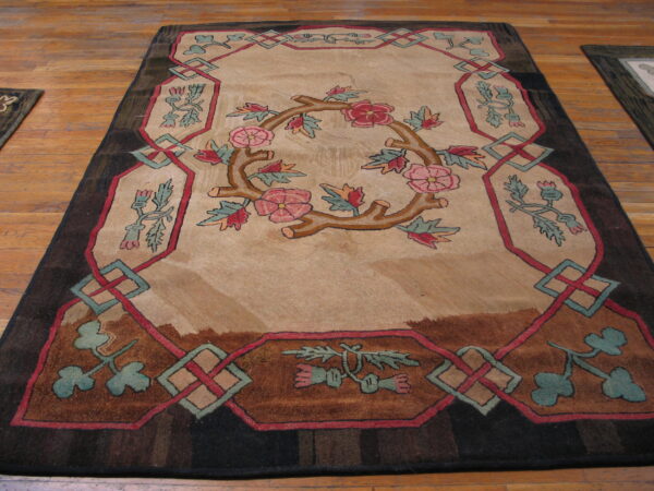 Tan rug with pink floral wreath, dark brown frame, and red-teal lattice border on hardwood flooring.