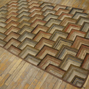 A textured rag rug with a chevron pattern in browns, tans, rust, and blue sits on distressed wood flooring.