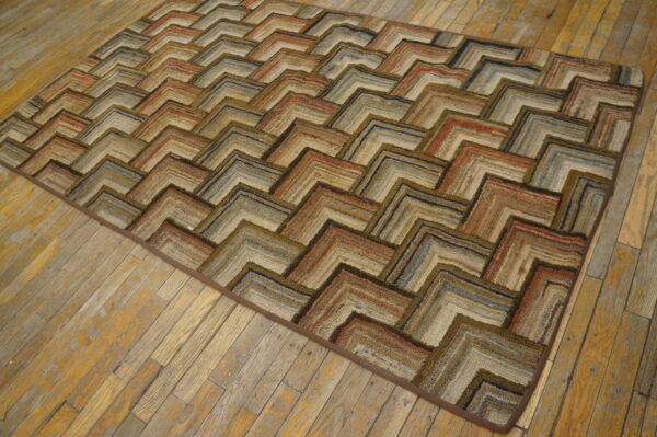 A textured rag rug with a chevron pattern in browns, tans, rust, and blue sits on distressed wood flooring.