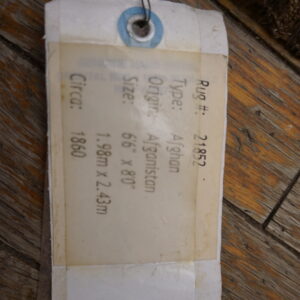 A white product identification tag rests on distressed brown wood planks beside a dark, high-pile rug edge.