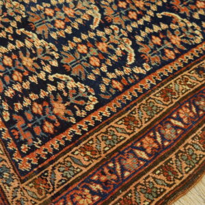 A dark blue rug with dense orange and peach repeating patterns, featuring multi-layered borders on light wood.