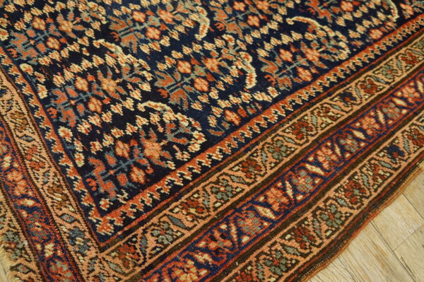 A dark blue rug with dense orange and peach repeating patterns, featuring multi-layered borders on light wood.
