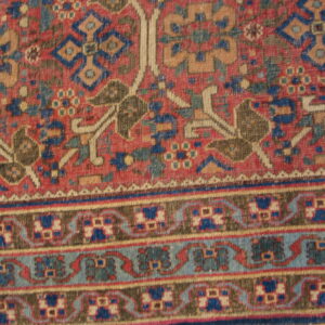 Detail of a traditional low-pile rug featuring a red field, blue and tan geometric motifs, and multiple borders.
