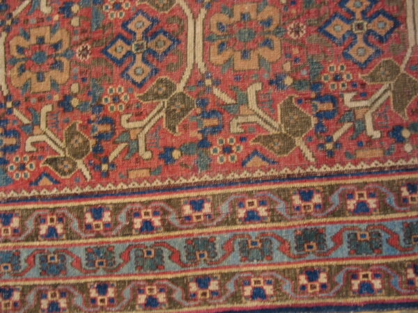 Detail of a traditional low-pile rug featuring a red field, blue and tan geometric motifs, and multiple borders.