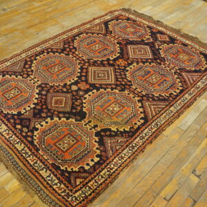 Low-pile geometric rug in dark blue, rust, and beige, displayed on worn yellow wood planks.