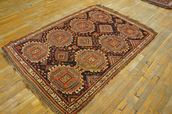 Low-pile geometric rug in dark blue, rust, and beige, displayed on worn yellow wood planks.