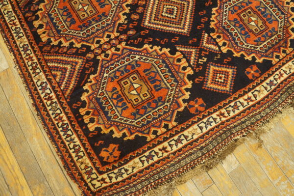 Corner view of a dark geometric rug with orange and blue medallions laid on light wooden floorboards.