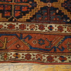 Detailed low-pile rug section with dense geometric patterns in rust red, navy blue, and gold resting on wood.