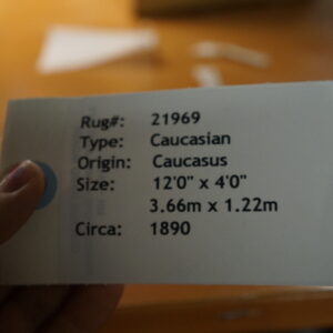 A hand holds a white identification tag with black text over a blurred warm, orange-brown surface.