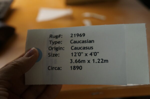 A hand holds a white identification tag with black text over a blurred warm, orange-brown surface.