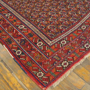 Red and blue geometric rug corner with detailed borders sits on distressed light brown wood flooring.