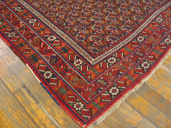 Red and blue geometric rug corner with detailed borders sits on distressed light brown wood flooring.