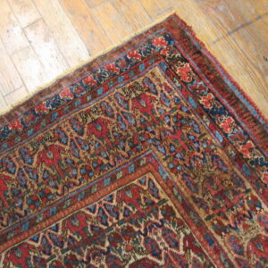 Low-pile red, navy, and tan traditional patterned rug corner on worn plank wood flooring.
