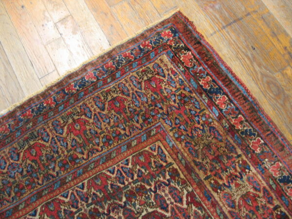 Low-pile red, navy, and tan traditional patterned rug corner on worn plank wood flooring.