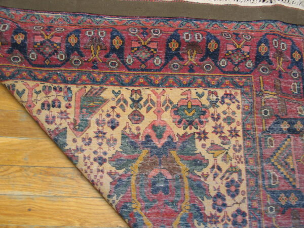 A colorful, low-pile traditional rug is folded on a wood floor, exposing its backing and intricate pattern.