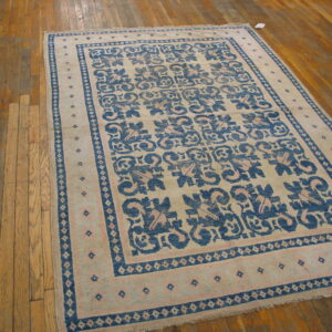 A beige rug with blue and pink geometric patterns and borders rests on worn plank wood flooring.