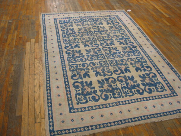 A beige rug with blue and pink geometric patterns and borders rests on worn plank wood flooring.