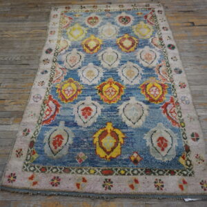 Textured rug with a blue field, colorful repeating motifs, and a light pink border on distressed wood flooring.