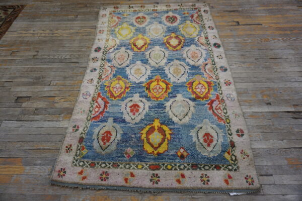 Textured rug with a blue field, colorful repeating motifs, and a light pink border on distressed wood flooring.