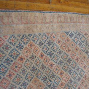 Beige rug with a diamond lattice field pattern in faded blue and pink on natural wood floor.