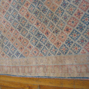 Geometric diamond pattern rug in faded blue, pink, and tan with a striped border, placed on wood floors.