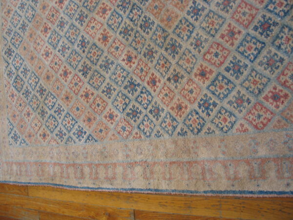 Geometric diamond pattern rug in faded blue, pink, and tan with a striped border, placed on wood floors.