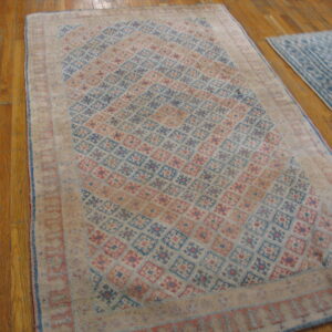 A distressed rug with blue and rose geometric diamonds sits on hardwood flooring next to a lighter blue rug.