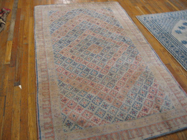 A distressed rug with blue and rose geometric diamonds sits on hardwood flooring next to a lighter blue rug.
