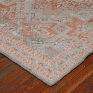 Corner of a textured blue and peach geometric rug lying on reddish-brown hardwood planks.