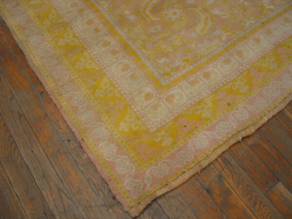 Corner view of a blush pink and yellow traditional rug with borders on plank wood flooring.