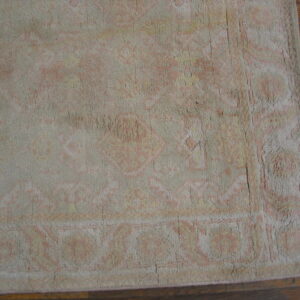 Close-up of a low-pile rug with faded pink, yellow, and beige traditional patterns on brown hardwood flooring.