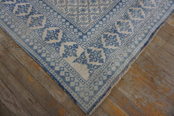 Faded blue and off-white rug with bordered patterns, showing tassels on distressed wood floor.