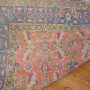 The corner of a low-pile rug with blue, coral, and green traditional patterns rests on wooden planks.
