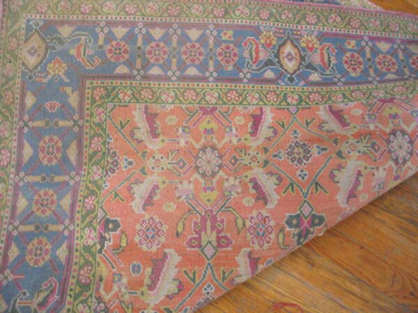 The corner of a low-pile rug with blue, coral, and green traditional patterns rests on wooden planks.