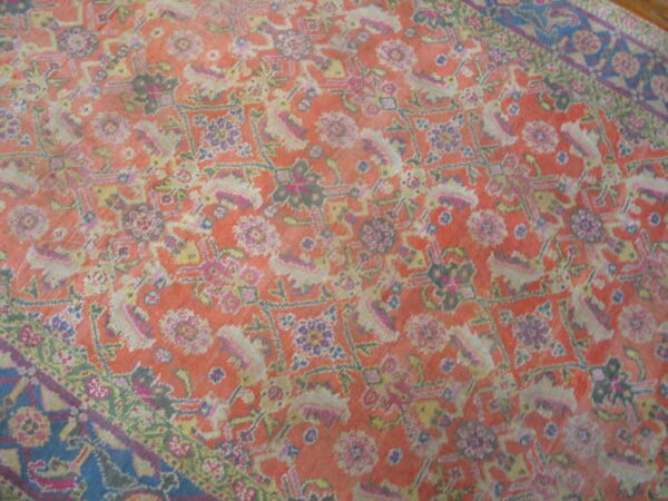 Distressed traditional rug featuring an orange-red field, ornate floral patterns, and a dark blue patterned border.