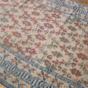 Tan low-pile rug with faded red and blue botanical patterns, showing visible seams on wood flooring.
