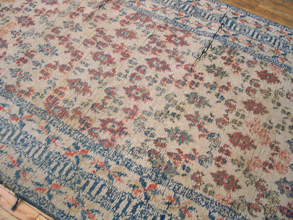 Tan low-pile rug with faded red and blue botanical patterns, showing visible seams on wood flooring.