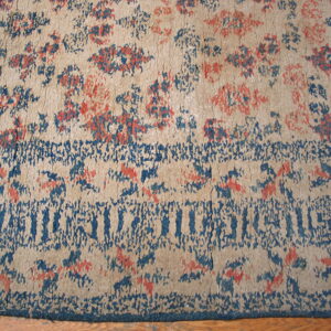 Worn low-pile rug in beige, deep blue, and coral featuring abstract floral motifs and a distressed border.