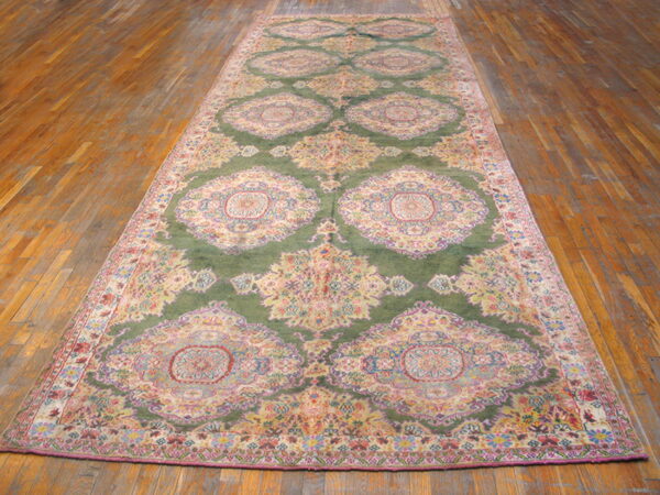 Long runner rug with repeating pink and cream medallions on a deep olive green field over wood floors.