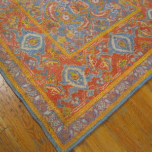 Faded traditional rug features coral, blue, and gold colors against worn medium-toned plank flooring.