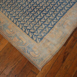 Blue and tan patterned rug corner with light border sits on dark, rustic wooden floor planks.