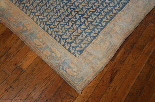 Blue and tan patterned rug corner with light border sits on dark, rustic wooden floor planks.
